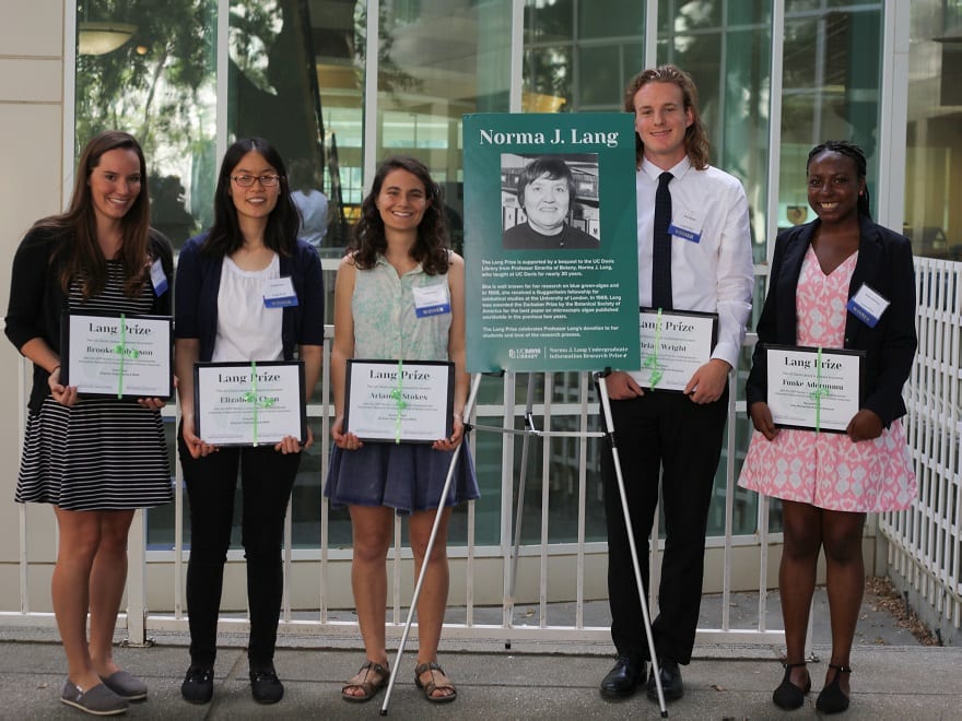 The 2017 Lang Prize winners stand holding their certificates, on either side of a large photo of the late Professor Emerita Norma J. Lang